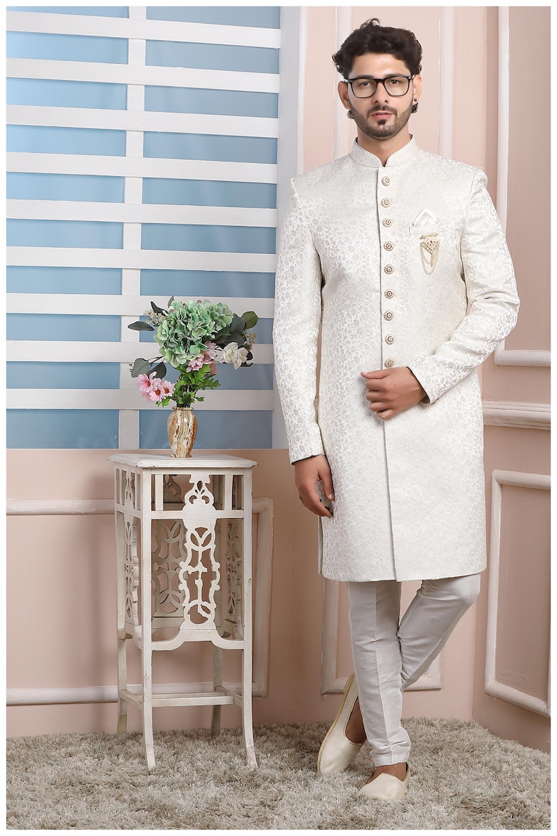 Puede incluir: Una kurta blanca de manga larga con cuello mandar&iacute;n y cierre de botones. La kurta tiene un sutil estampado floral y se combina con pantalones blancos. El hombre lleva zapatos blancos y gafas.