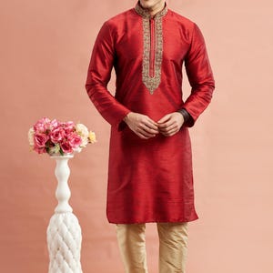 May include: A man wearing a red kurta with gold embroidery around the collar and down the front. He is also wearing gold-colored pants and white shoes. A white decorative vase with pink flowers is to the left.