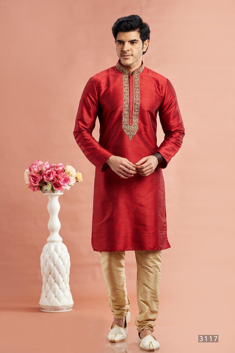 May include: A man wearing a red kurta with gold embroidery around the collar and down the front. He is also wearing gold-colored pants and white shoes. A white decorative vase with pink flowers is to the left.