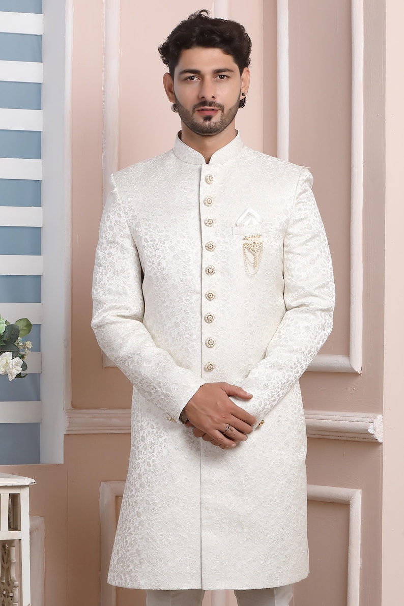 Puede incluir: Una camisa blanca de manga larga con botones, un patr&oacute;n texturizado y botones dorados. La camisa tiene un cuello mandar&iacute;n y un bolsillo en el lado izquierdo con una cadena dorada y una pieza decorativa.