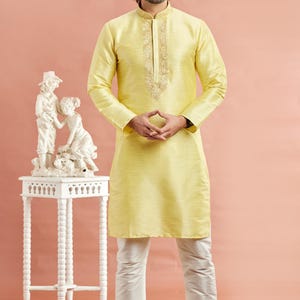 May include: A man wearing a yellow kurta with intricate embroidery around the collar, paired with white pants and cream-colored shoes. A white decorative table with a statue is to the left. The background is a solid peach color.