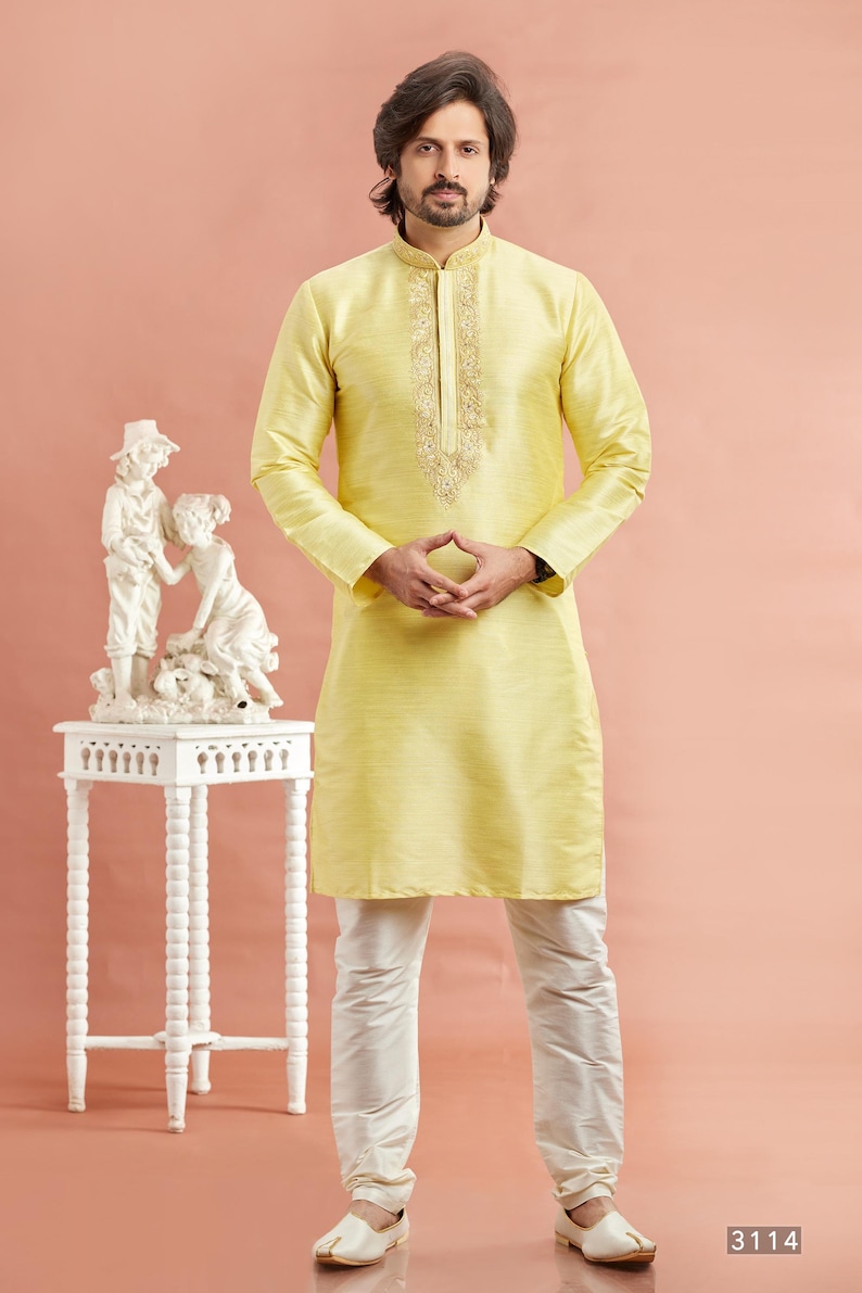 May include: A man wearing a yellow kurta with intricate embroidery around the collar, paired with white pants and cream-colored shoes. A white decorative table with a statue is to the left. The background is a solid peach color.