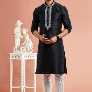 May include: Black kurta with white embroidery on the collar and a matching pair of white pants. The long-sleeved kurta is paired with white shoes. The outfit is worn by a person wearing sunglasses.