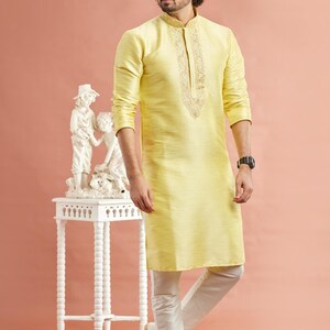 May include: A man wearing a light yellow kurta with intricate embroidery around the collar and neckline, paired with white pants and shoes. The outfit is traditional Indian attire. A small white table with a sculpture is to the left.