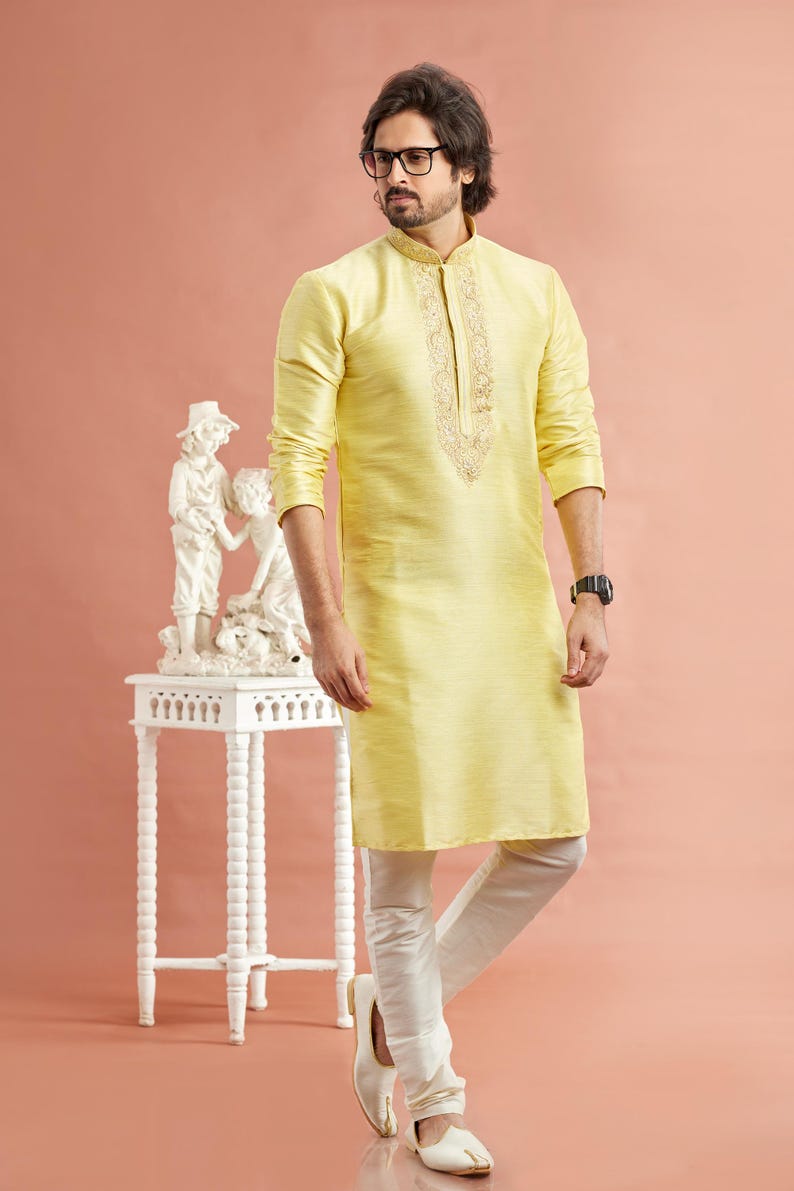 May include: A man wearing a light yellow kurta with intricate embroidery around the collar and neckline, paired with white pants and shoes. The outfit is traditional Indian attire. A small white table with a sculpture is to the left.