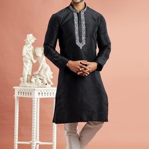 May include: A black kurta with white embroidery around the collar and neckline, paired with white pants. The long-sleeved tunic has a straight cut. The outfit is worn with white shoes. The image is set against a peach background.