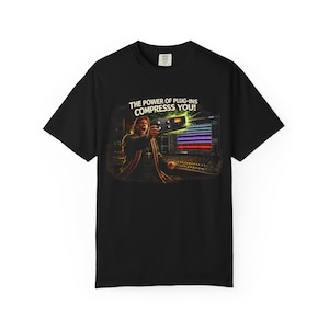 May include: Black t-shirt featuring a graphic of a person in a robe pointing a device at a sound mixing board. The text "THE POWER OF PLUG-INS COMPRESSSS YOU!" is above the graphic. The shirt is made of a soft material.