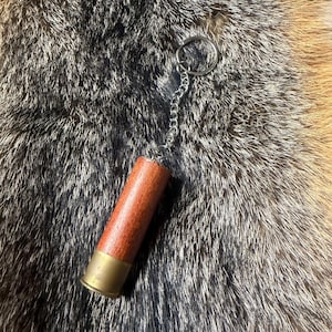 Puede incluir: Un llavero de cartucho de escopeta con base de latón y cuerpo de madera. El llavero está sujeto a una cadena y un anillo plateados. El fondo es una piel texturizada.