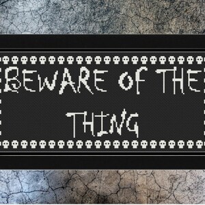 以下が含まれることがあります： 黒地に白の文字で「BEWARE OF THE THING」と書かれた、黒枠で囲まれた白黒のクロスステッチデザインです。