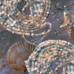 Puede incluir: Primer plano de joyería hecha a mano con temática marina. El diseño presenta tela azul enrollada adornada con cuentas blancas, azules y plateadas, junto con perlas de imitación y detalles de cristal transparente. Se ven conchas marinas y estrellas de mar en el fondo.