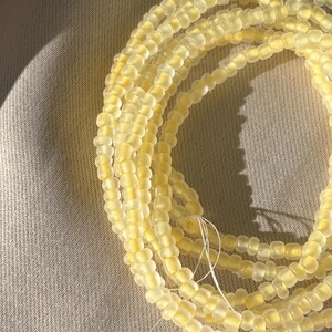 Puede incluir: Un hilo enrollado de cuentas de cintura de color amarillo pálido. Las cuentas son pequeñas, cilíndricas y están ensartadas en un hilo blanco fino. Las cuentas tienen un aspecto ligeramente translúcido, y el fondo es una tela neutra y texturizada.