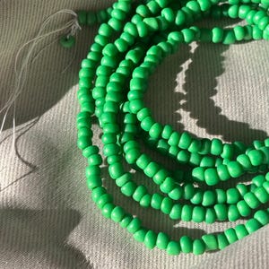Puede incluir: Varias hebras de cuentas verdes brillantes, probablemente para la fabricación de joyas o manualidades, están dispuestas en espiral. Las cuentas, pequeñas y redondas, son de color uniforme. Se ve un hilo blanco en el extremo, lo que indica que están ensartadas.