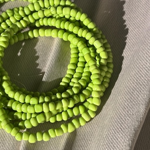 Puede incluir: Collar de cuentas verde brillante enrollado sobre una superficie gris claro texturizada. Las pequeñas cuentas cilíndricas están espaciadas uniformemente, creando un accesorio llamativo. Probablemente una joya corporal.