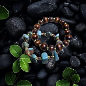 Puede incluir: Dos pulseras de cuentas. Una pulsera presenta grandes cuentas redondas de madera marrón oscuro. La otra pulsera tiene cuentas de piedra turquesa y marrón. Ambas pulseras tienen espaciadores dorados y se exhiben sobre piedras negras.