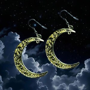 Puede incluir: Un par de pendientes dorados con forma de media luna y detallados diseños de filigrana. Los pendientes tienen cierre de gancho. El fondo es oscuro con nubes y estrellas, creando un tema celestial. Miden aproximadamente 3 cm.