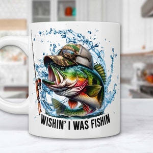 Könnte beinhalten: Weiße Keramik-Kaffeetasse mit einer bunten Illustration eines Barsches, der aus dem Wasser springt. Der Fisch trägt einen Tarnhut. Der Text "Wishin' I was fishin'" ist auf die Tasse gedruckt.