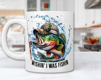 Puede incluir: Taza de café de cerámica blanca con una ilustración colorida de una lubina saltando fuera del agua. El pez lleva un sombrero de camuflaje. El texto "Wishin' I was fishin'" está impreso en la taza.