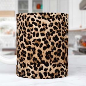 Pode incluir: Uma caneca de café de cerâmica branca com um design de estampa de leopardo. A caneca tem um padrão marrom, preto e bege.