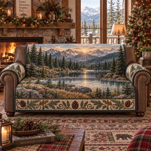 May include: A sofa cover featuring a landscape print of a mountain range, a lake, and a pine forest at sunset. The fabric has a quilted texture and a decorative border along the bottom edge with pinecone and evergreen branch illustrations.