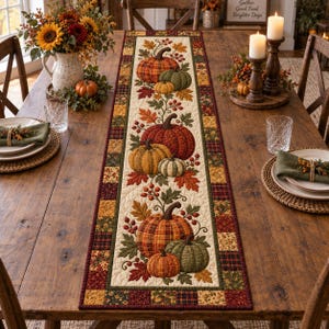 Puede incluir: Un camino de mesa acolchado rectangular con un panel central de color crema decorado con un patrón repetitivo de calabazas naranjas, rojas y verdes, hojas de otoño y bayas, bordeado por un mosaico de cuadrados de tela a cuadros y florales.
