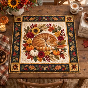 May include: A square fabric mat featuring a central illustration of a sleeping orange tabby cat curled within a wreath of autumn leaves, sunflowers, red flowers, wheat, and small pumpkins. The border has a dark frame with acorn motifs.