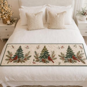 May include: A rectangular quilted table runner featuring a cream-coloured background with a repeating pattern of green pine trees, red cardinals, pinecones, and holly berries, finished with a dark green border along the edges of the fabric.