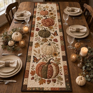 Peut inclure: Un chemin de table matelassé rectangulaire avec un panneau central crème orné d'un arrangement vertical de citrouilles à motifs variés, de feuilles d'automne, de tournesols et de papillons, bordé d'une garniture marron foncé et à carreaux.