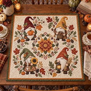 May include: A square fabric quilt featuring four gnomes in autumn-themed attire, each holding seasonal items like a pumpkin, a basket of acorns, a sunflower, and a lantern, surrounded by embroidered maple leaves, acorns, and a central flower.