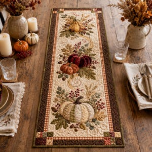 May include: A rectangular quilted table runner featuring a cream-coloured centre panel decorated with embroidered pumpkins, autumn leaves, and berries. The runner has a dark brown border with a small checkered pattern and intricate stitching throughout.