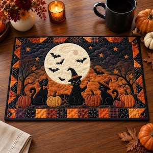 May include: A rectangular quilted placemat featuring a Halloween scene with three black cats, one wearing a witch hat and holding a wand, set against a large full moon, bats, orange pumpkins, and bare trees, framed by a patchwork border of autumn colors.