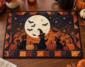 May include: A rectangular quilted placemat featuring a Halloween scene with three black cats, one wearing a witch hat and holding a wand, set against a large full moon, bats, orange pumpkins, and bare trees, framed by a patchwork border of autumn colors.
