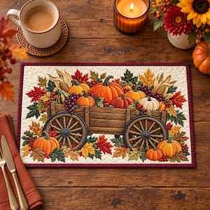 Può includere: Una tovaglietta trapuntata rettangolare con un'illustrazione centrale di un carro in legno colmo di zucche arancioni, zucche ornamentali e foglie autunnali nei toni del rosso, giallo e verde. Il design è rifinito da un bordo in tessuto rosso scuro.