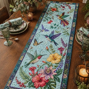 Pode incluir: Um caminho de mesa retangular acolchoado com fundo azul-claro e uma estampa vibrante de beija-flores, borboletas e várias flores tropicais, incluindo hibiscos e flores-da-paixão, finalizado com uma borda escura estampada.