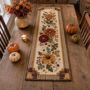 May include: A rectangular fabric table runner featuring a cream-colored center panel with a printed design of orange, red, and yellow sunflowers, autumn leaves, and berries, bordered by a brown and tan checkered pattern and dark green trim.