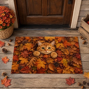 May include: A rectangular doormat featuring a high-resolution print of an orange tabby cat peeking out from a dense pile of fallen autumn leaves in shades of red, orange, yellow, and brown, with the cat's paws resting on the foliage.