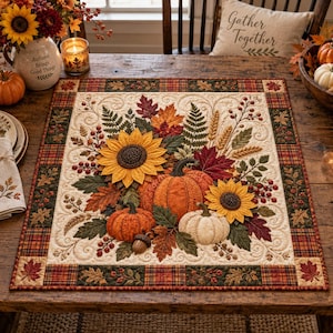 May include: A square fabric table mat featuring a quilted autumn design with two yellow sunflowers, an orange pumpkin, a white pumpkin, an acorn, and various fall leaves, berries, and wheat stalks, framed by a plaid border in red, green, and brown.