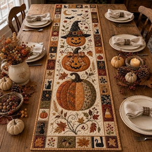 May include: A rectangular fabric table runner featuring a patchwork border and a central design of three stacked pumpkins, one wearing a witch hat, surrounded by autumn leaves, vines, black cats, and bats, placed on a wooden dining table.