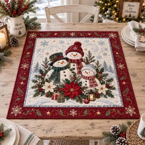 May include: A rectangular quilted table mat featuring three snowmen in a snowy landscape with poinsettias, pine branches, and small gift boxes. The border is deep red with gold snowflake and holly leaf embroidery on a cream-coloured background.