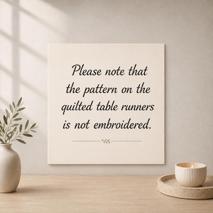 May include: A square off-white sign with black text that reads: Please note that the pattern on the quilted table runners is not embroidered. Below the text is a small horizontal line with a decorative leaf icon in the center.