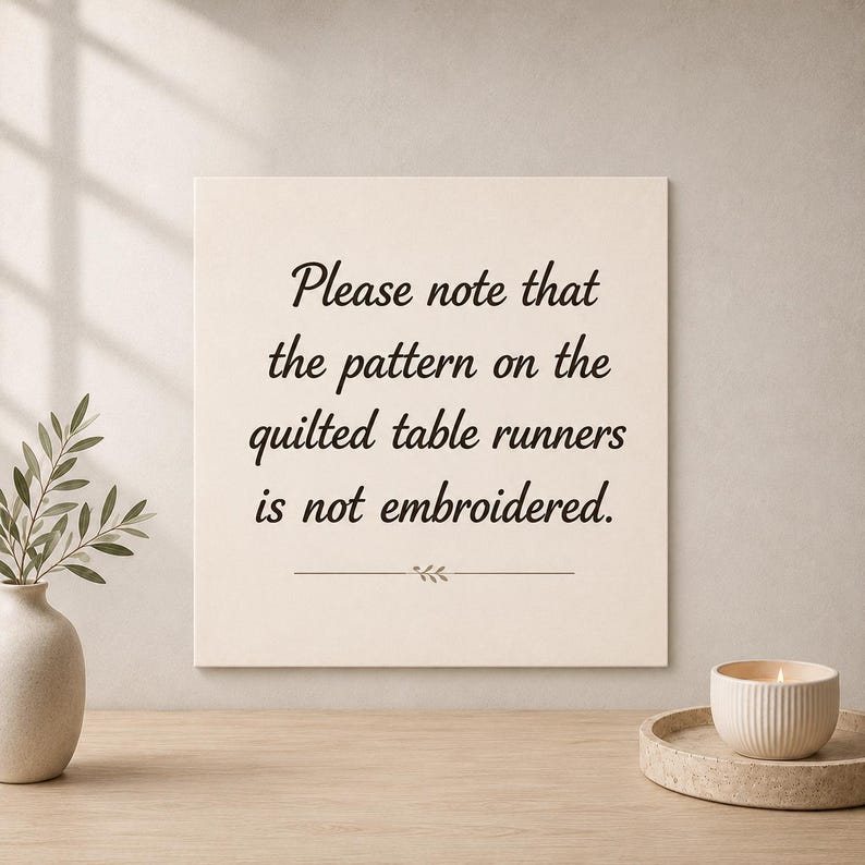 May include: A square off-white sign with black text that reads: Please note that the pattern on the quilted table runners is not embroidered. Below the text is a small horizontal line with a decorative leaf icon in the center.
