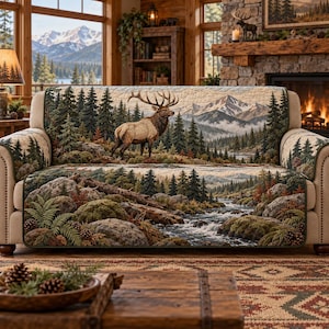 May include: A quilted sofa cover featuring a detailed landscape print of a majestic elk standing in a pine forest near a flowing river, with snow-capped mountains in the background. The design includes mossy rocks, ferns, and pinecones.
