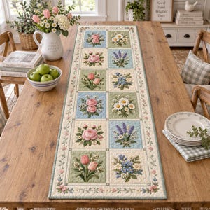 Puede incluir: Un camino de mesa acolchado rectangular que presenta una cuadrícula de cuadrados florales con representaciones bordadas de rosas rosas, tulipanes, lavanda y margaritas. El camino tiene un borde color crema con un delicado patrón de enredaderas.