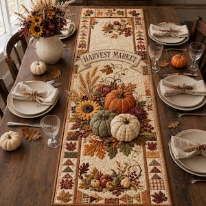 Peut inclure: Un chemin de table matelassé rectangulaire orné d'une bannière Harvest Market, de citrouilles, de tournesols et de feuilles d'automne dans des tons orange, vert et marron. Il est posé sur une table en bois avec des couverts et des serviettes.