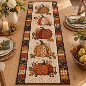 May include: A rectangular quilted table runner featuring a central cream-coloured panel with five appliqued fabric pumpkins in shades of orange, brown, and green, surrounded by a border of small, multi-coloured patchwork squares in autumn tones.