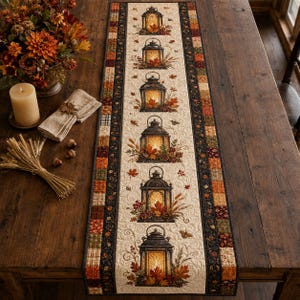 May include: A rectangular quilted table runner featuring a central cream-colored panel decorated with a vertical row of six black lanterns surrounded by autumn leaves, pinecones, and berries, framed by dark borders with colorful patchwork squares.