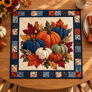 May include: A square fabric table mat featuring a central quilted design of pumpkins in orange, blue, green, and white, surrounded by autumn leaves, wheat stalks, and acorns. The border consists of a patchwork pattern in navy blue and floral prints.
