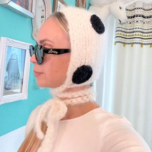 Könnte beinhalten: Eine weiße Strickmütze mit zwei großen schwarzen Punkten und einem passenden Schal, der um den Hals gebunden ist. Die Mütze wird mit einer schwarzen Sonnenbrille mit dem Wort "Loewe" auf der Seite getragen.