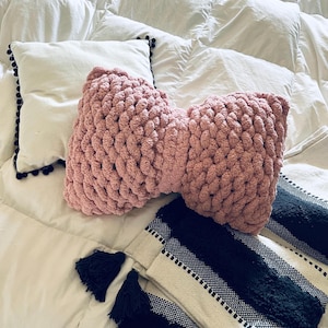 May include: A pink knitted bow pillow on a white comforter with a black and white striped throw blanket with tassels