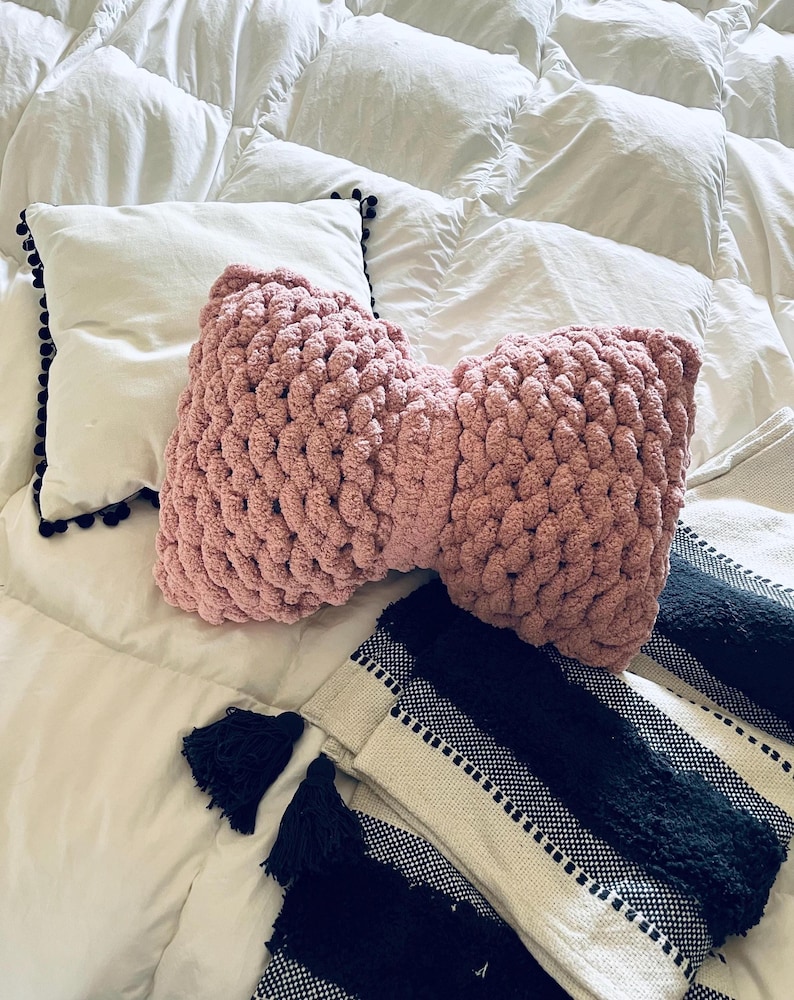 May include: A pink knitted bow pillow on a white comforter with a black and white striped throw blanket with tassels