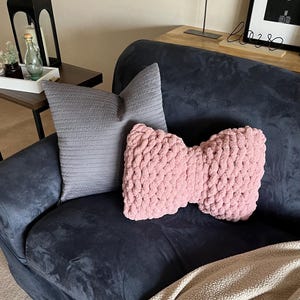 May include: A navy blue sofa with two decorative pillows and a blanket. One pillow is a light gray, rectangular shape. The other is a pink, bow-shaped, chunky knit pillow. A beige blanket is draped over the sofa.